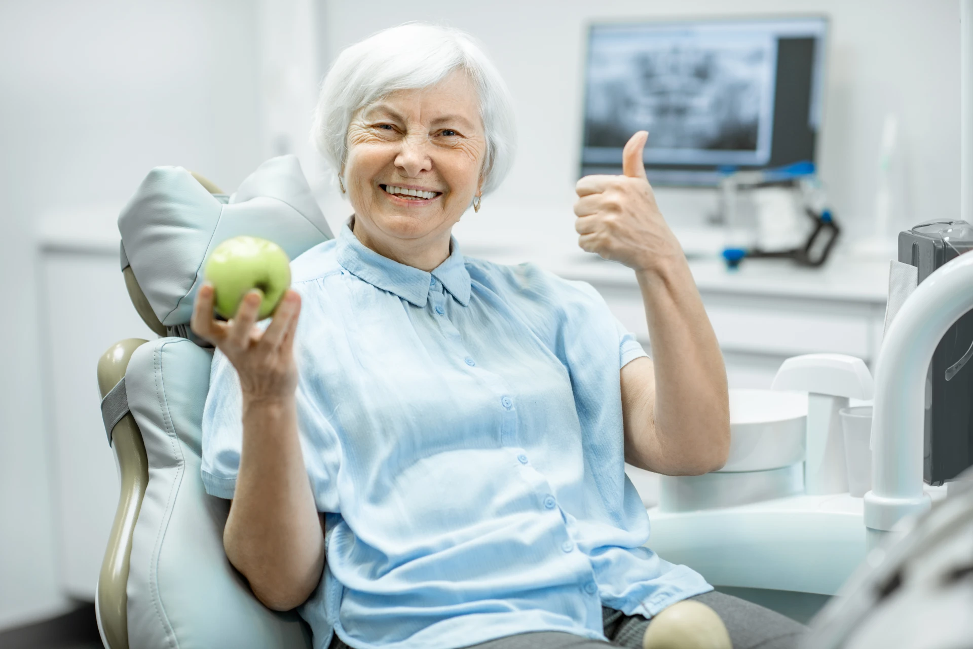 Photo of a senior woman in the dental chair smiling while holding an apple and giving a thumbs up.